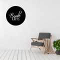 Beach Life coastal typography metal wall art displayed above a modern accent chair, featuring elegant script lettering inside a circular laser-cut steel design.