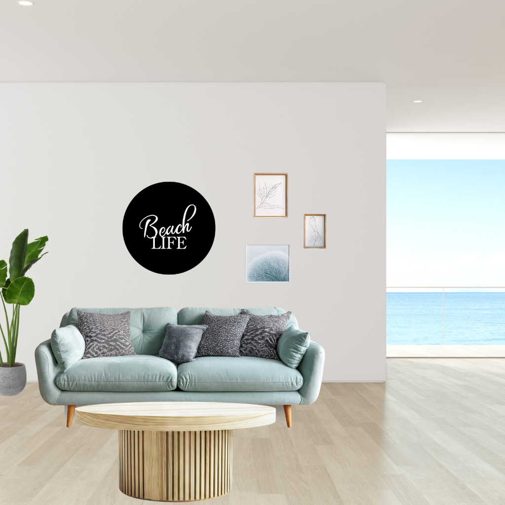 Beach Life circular metal wall art displayed above a contemporary sofa in a bright coastal-inspired living room overlooking the ocean.