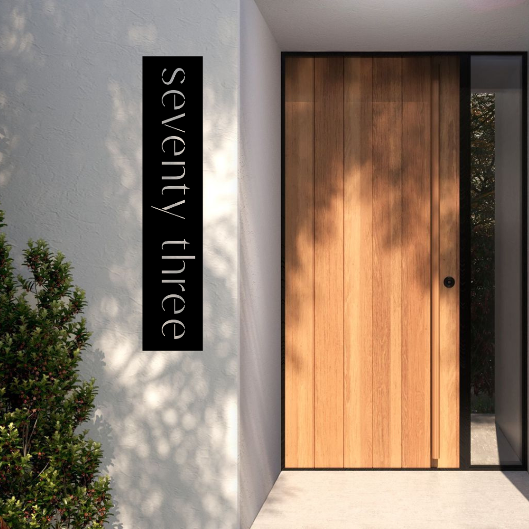 Minimalist vertical metal address sign installed on a modern rendered entrance wall beside a timber front door.