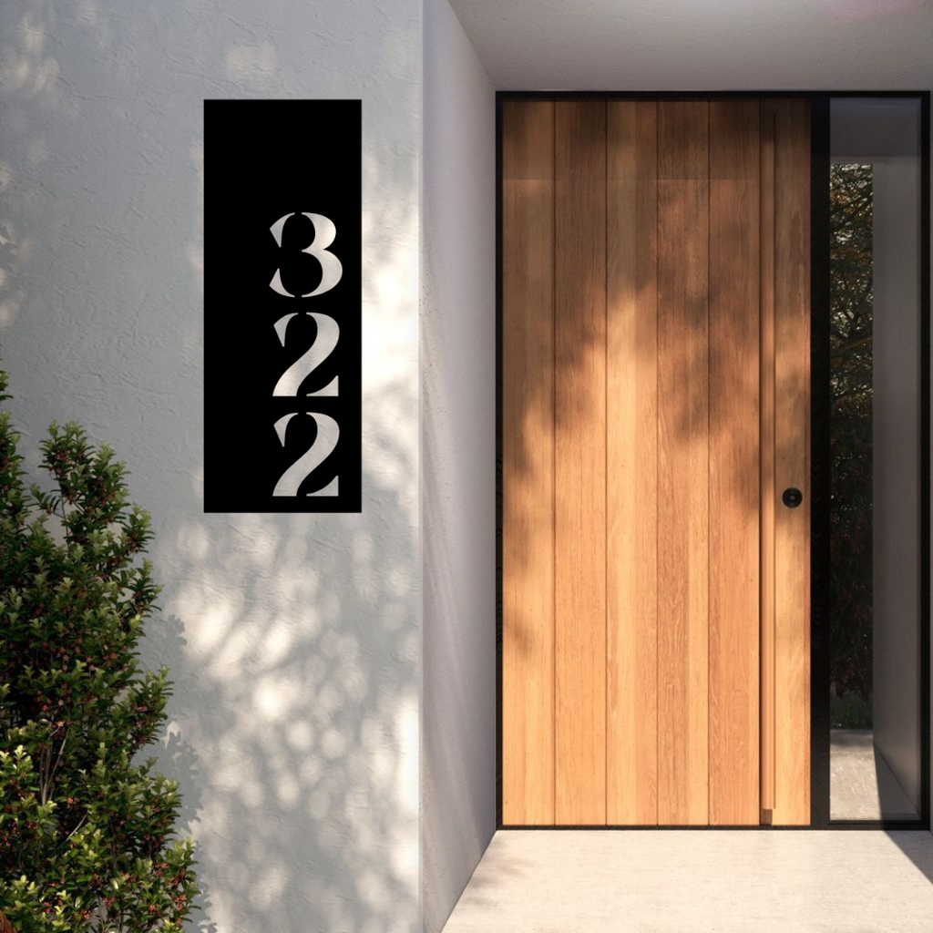 Modern vertical metal house number installed on a rendered entrance wall beside a contemporary timber front door.